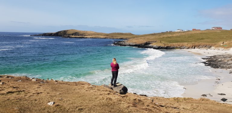 Figure of woman in pink top standing on edge of cliff looking over bay with white sand and turquoise sea