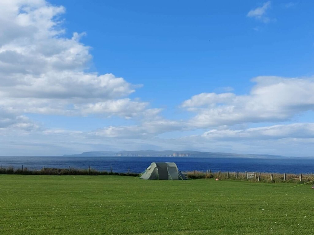 View of green field, green tent at edge of cliff and blue see and sky 