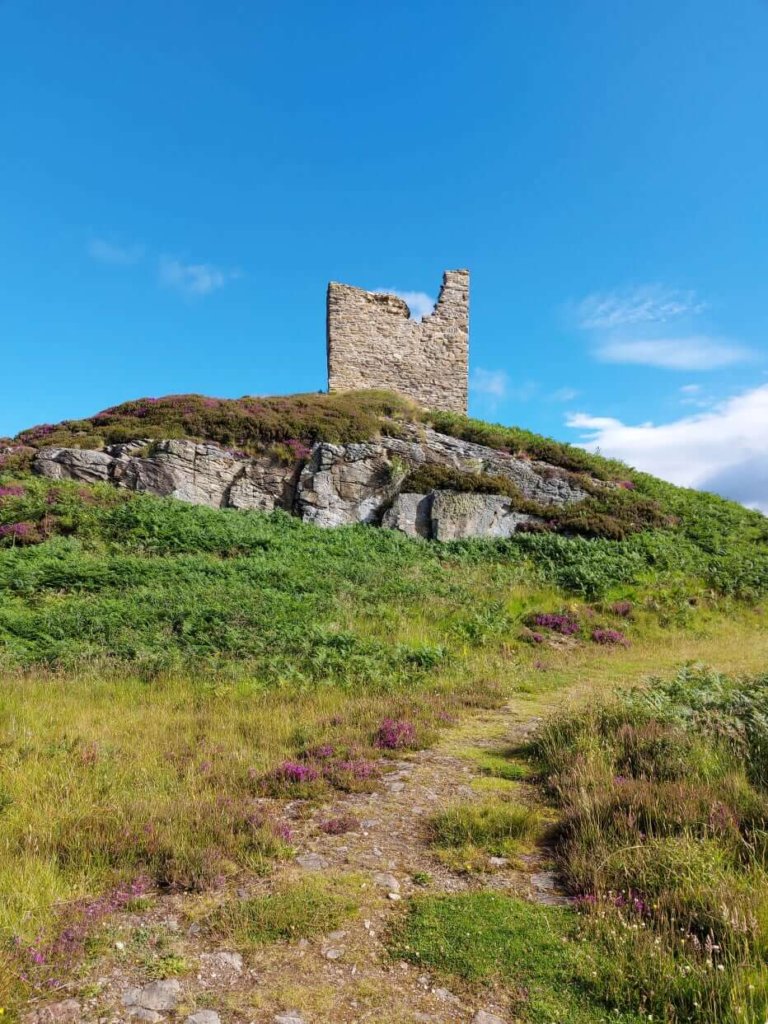 Solo female travel
Ruin of castle on top of hill with green grass, purple heather and rocks, blue sky background