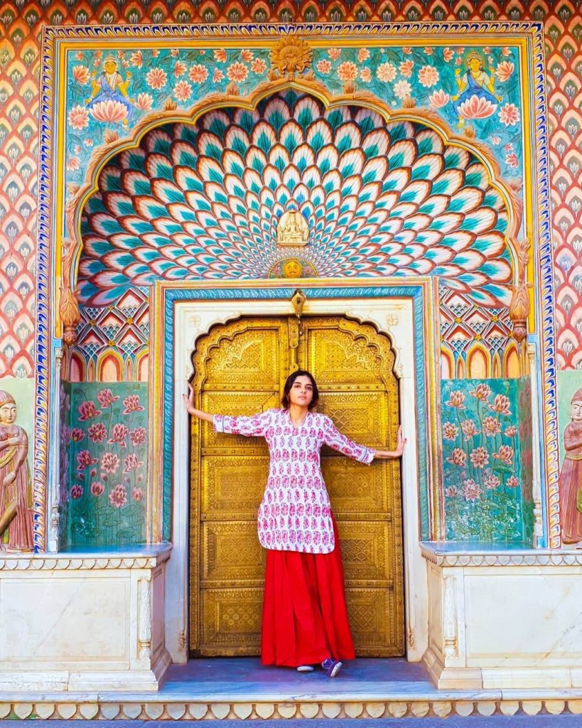 Solo female travel in India 
Woman with short black ahir wearing long red skirt and pink and white top, standing in fron of ornate door, with detailed painted surround in multi colours