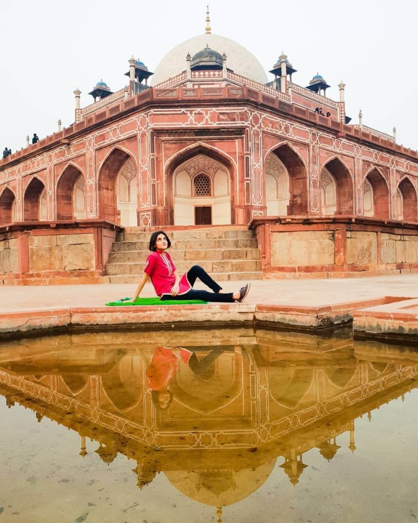 Solo female travel in India 
Woman with short black hair sat on green blanket in front of red Indian temple at Humayun's Tomb, pond in front reflecting image