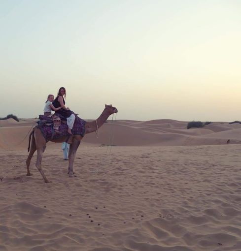 Woman and child sat on camel in desert hills at sunset