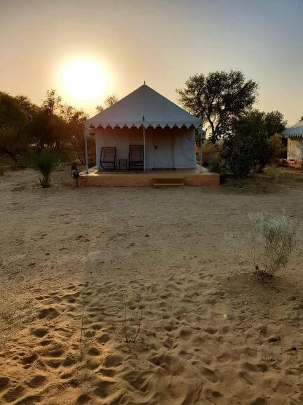 Desert camp circular tent with 2 chairs on small deck, sandy ground with small trees and shrubs Sun in hazy background