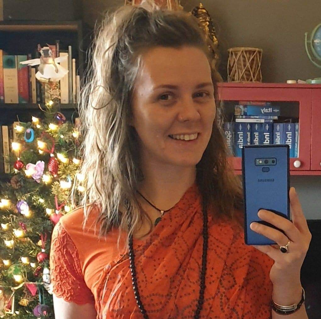 Solo female travel in India 
Woman with shoulder length blonde hair tied up loosely wearing orange sari and dark beaded necklace, Christmas tree in background lit with coloured lights and bookshelf with lonely planet guides