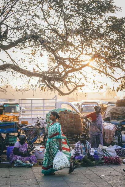 Indian woman carrying shopping walking past Indian street sellers with tree branches overhead and sun behind