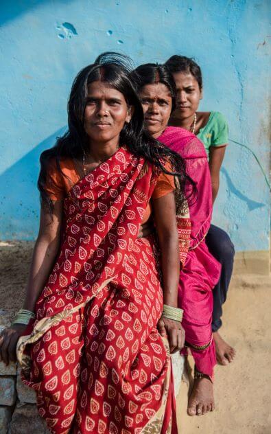 3 Indian women sat behind each other, wearing traditional sari in red patterned fabric, bright pink and green.  All the women have black hair