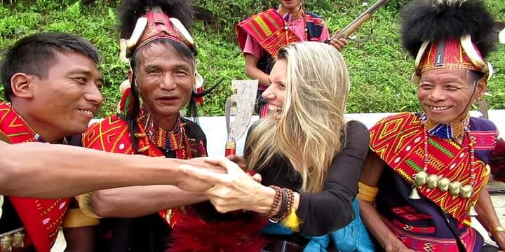 Blonde woman laughing with traditional dressed men of Konyak Tribe wearing red and bright coloured woven sashes and helmets with black fur