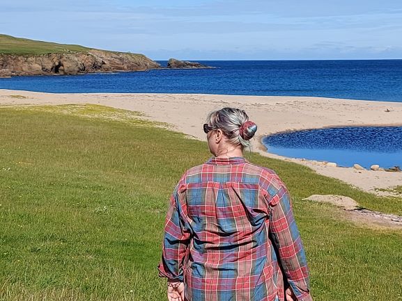 Travel trends solo female travel woman wearing red and blue checked shirt, from behind with green field, sandy beach and blue sea and sky in background