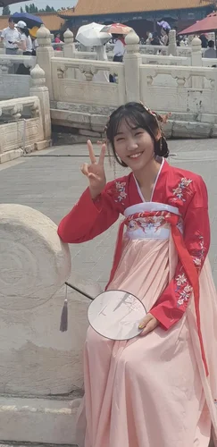 Chinese young woman with short black bob hair, in traditional red and peach outfit holding round fan on stick with tassel, smiling and giving peace sign