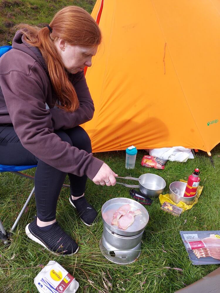 Woman sat on camp chair wearing purple hoodie, black legging and trainers cooking over camping stove with orange tent in background