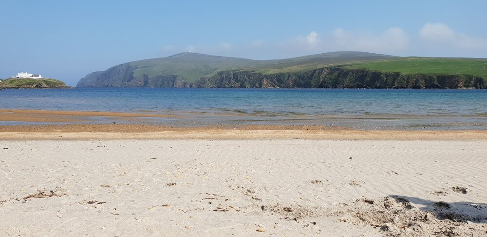 White sand beach, blue sea, cliffs topped by green fields in distance 