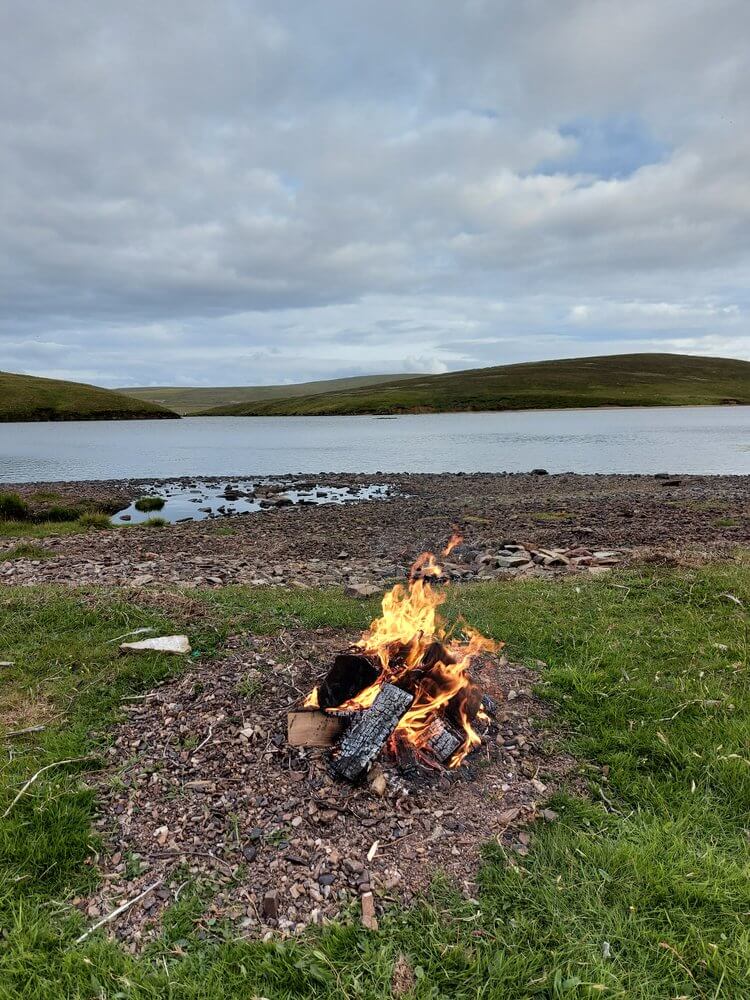 Campfire with orange and yellow flames in circle of stones with sea and hills in backgrouns