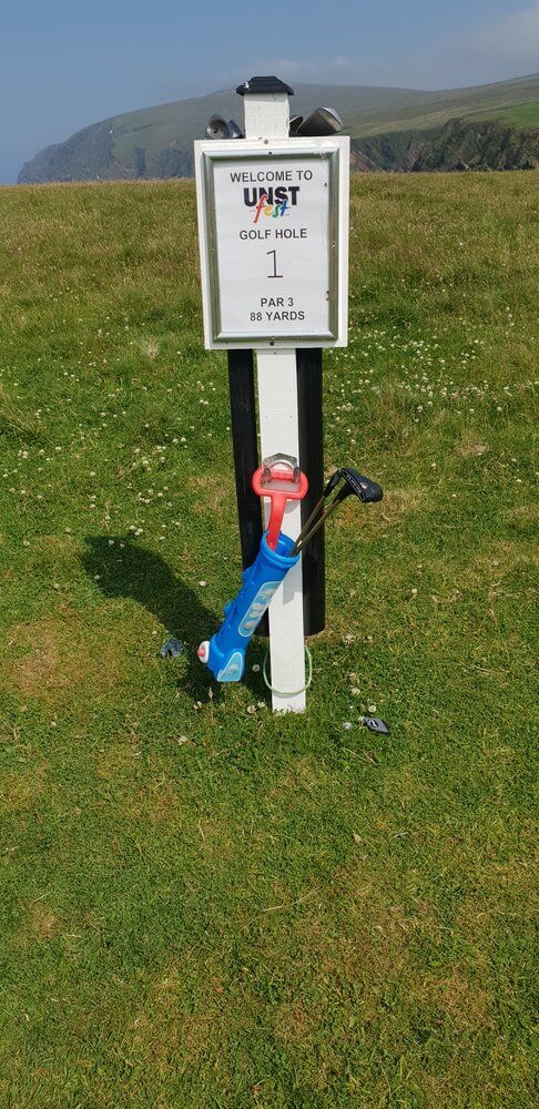 Child's bue and red plastic golf set leaning agianst a white wooden post with sign saying Welcome to Unst fest golf hole 1 par 3 88yards against green grass background