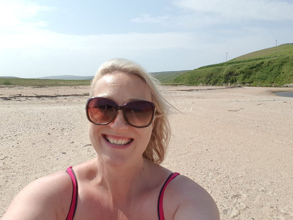 Head and shoulder shot of blonde woman sat on beach, smiling, wearing sunglasses, beach is the background 