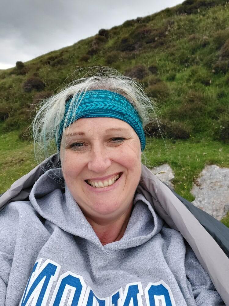 Selphie of woman wearing grey hoodie and turquoise bandana, with blonde hair sticking out of the top, green hill in background