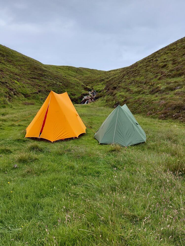 Orange and green tents in green field with green heather covered hill behind, grey sky