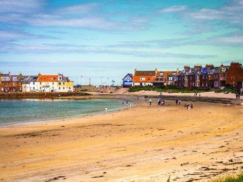 In search of beaches 
Curving beach in a bay, with houses on the right and in distance.  Some small figures of people in the distance, beach is empty in the foreground