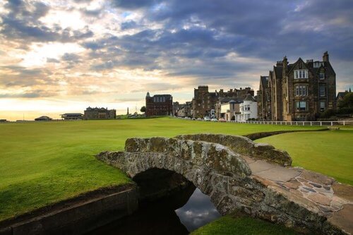 Day Trips from Edinburgh. Green golf course with small stone bridge over stream with grand clubhouse in background.  Cloudy sky at sunset