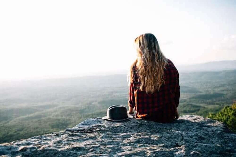 Travel Books
Woman sat high on a hill on a rocky outcrop looking out over the view of trees below.  She has long blonde hair, is wearing a red checked shirt and has a hat beside her