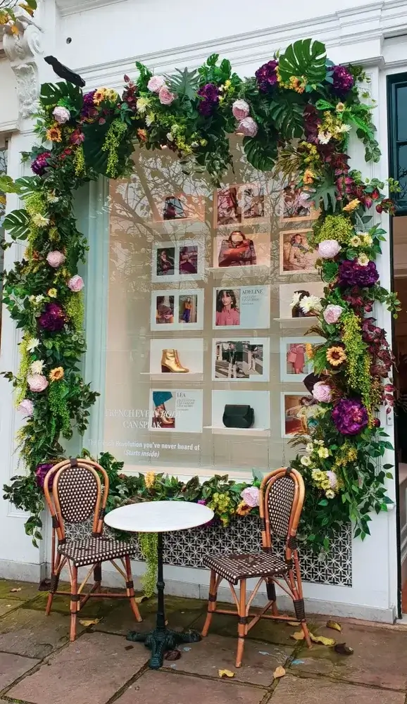 London
Shop window framed by real flowers, purple , pinks and yellow with green foliage, small round table with 2 chairs to front, window displaying a number of handbags and boots