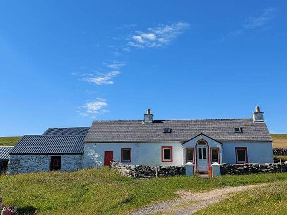 Road trip Shetland
White cottage with red window frames, 2 chimneys, outhouse to left, grass to front with stony track leading to it.  Clear blue sky behind