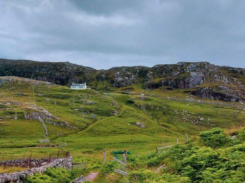 Solo female travel
White croft house, single storey with large windows in distance amidst rolling green hills with backdrop of rocky outcrops