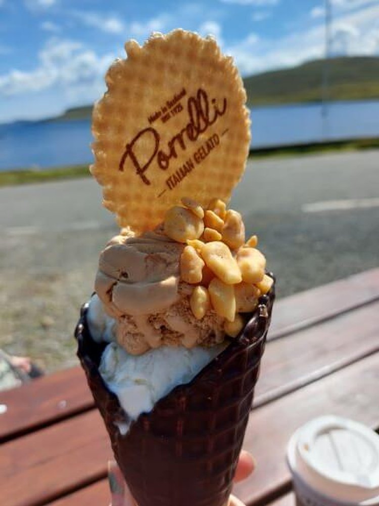 Fingers holding chocolate covered waffle cone with 2 kinds of ice cream, nuts and a wafer. picnic table, with takeaway cup and sea across road in background