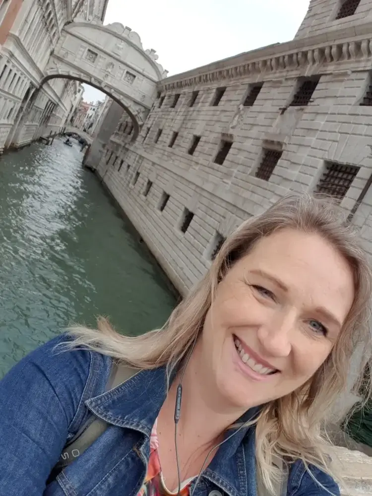 Woman standing in front of canal in venice with bridge of sighs behind her, she is blonde and wearing a denim jacket