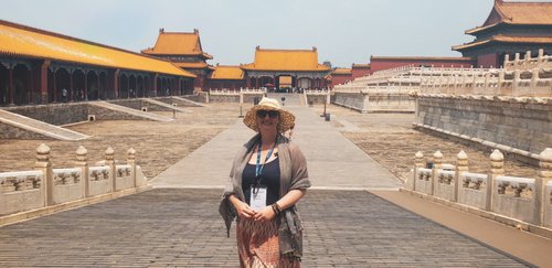 China
Woman in straw hat and sunglasses with grey shawl standing in empty  street of the Forbidden city, Chinese roofed building on either side, orange roofs with decorative edges and in distance