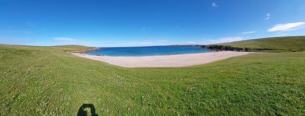 Plan a Stunning Road Trip to Unst, Shetland 
Green field leading to sandy beach and sea in a bay, clear blue sky