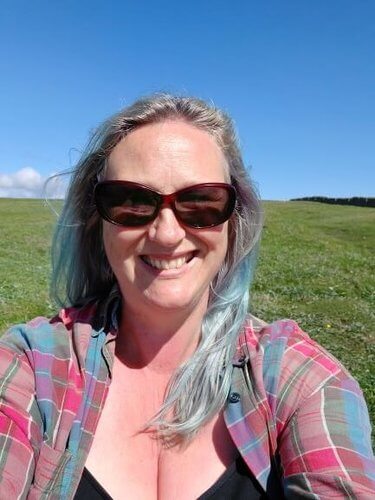 Woman with light blue hair, long, wearing sunglasses and black vest top, red and blue checked shirt against green field and blue sky