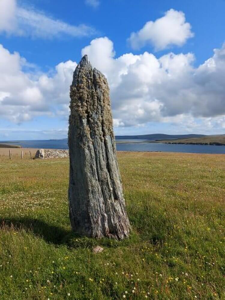 Large standing stone, weathered, standing in grass field with sea in distance blue sky with white clouds