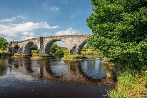 Stone bridge with 4 arches over river showing reflection, blue sky and green trees at right