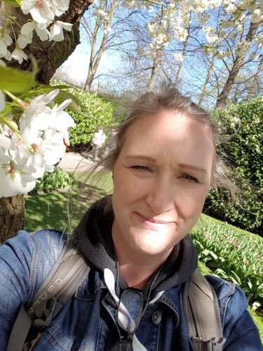 Selphie of woman in denim jacket carrying rucksack next to blossom tree with background of green bushes