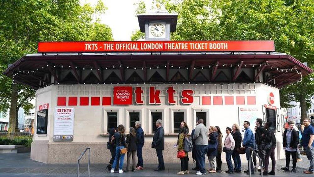 1 Things that will make you fall in love with London
TKTS booth in Leicester Square, with queue of people waiting, 4 windows, red trimming, clock tower on roof, trees behind