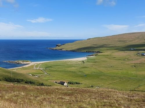 Looking down from hilltop over green fields to strip of white sand and deep blue sea and blue sky