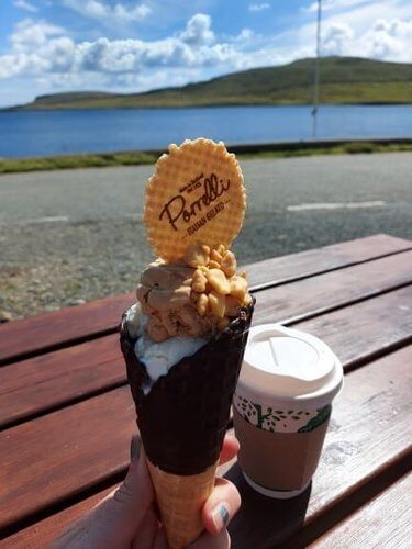 Fingers holding chocolate waffle cone with ice cream, nuts and wafer, takeaway coffee cup on brown wood table, sea and green hills in distance over road