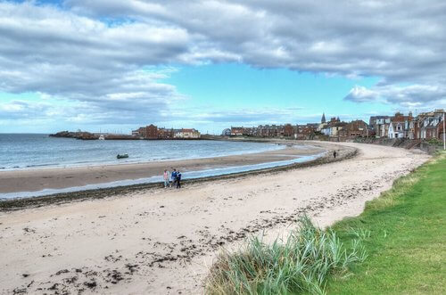 White sandy beach edged with grass, blue sea in a curved bay with house along edge in distance