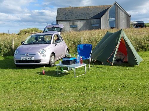 Solo female travel camping
Pink fiat 500 with boot open next to green tent, camping chair and table, wooden house behind, in green mown field