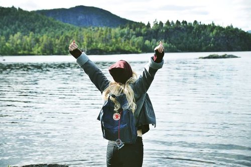 Woman standing by lake taken from behind, she is raising her arms in celebration. She is wearing a rucksack and a wooly hat