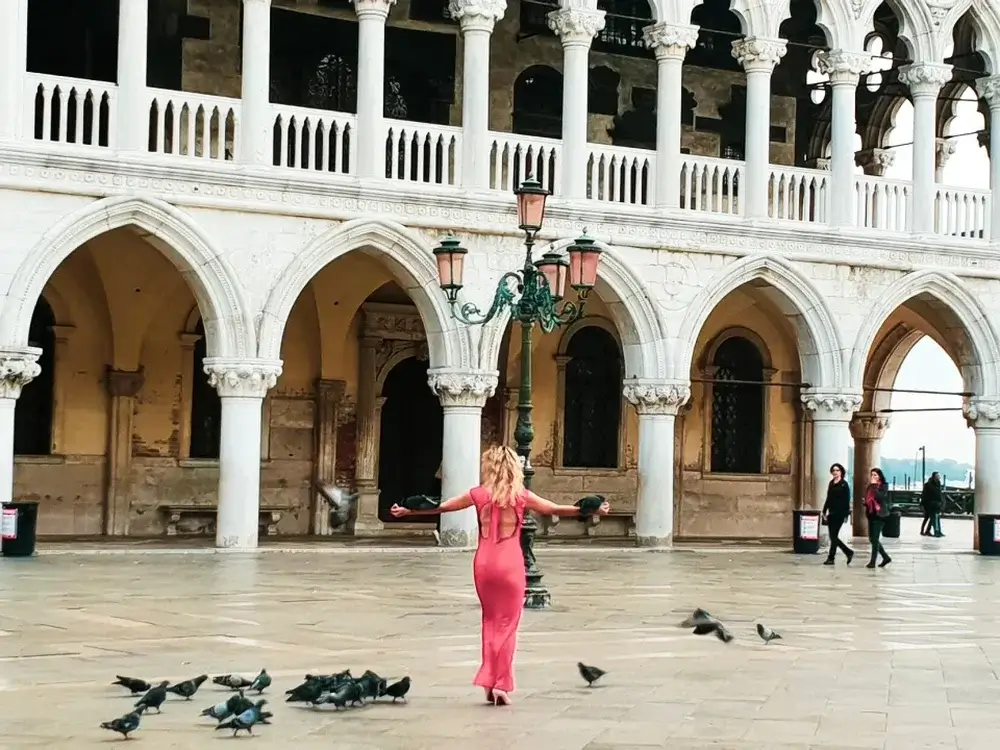 Being a tourist in Venice
Woman with arms out to the side in long pink dress surrounded by pigeons in front of arched walkway of Doges Palace