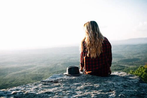 China
Woman with long blonde hair, in red checked shirt, sat on rocky outcrop at top of mountain looking down over view of forest.  Hat beside her on rock