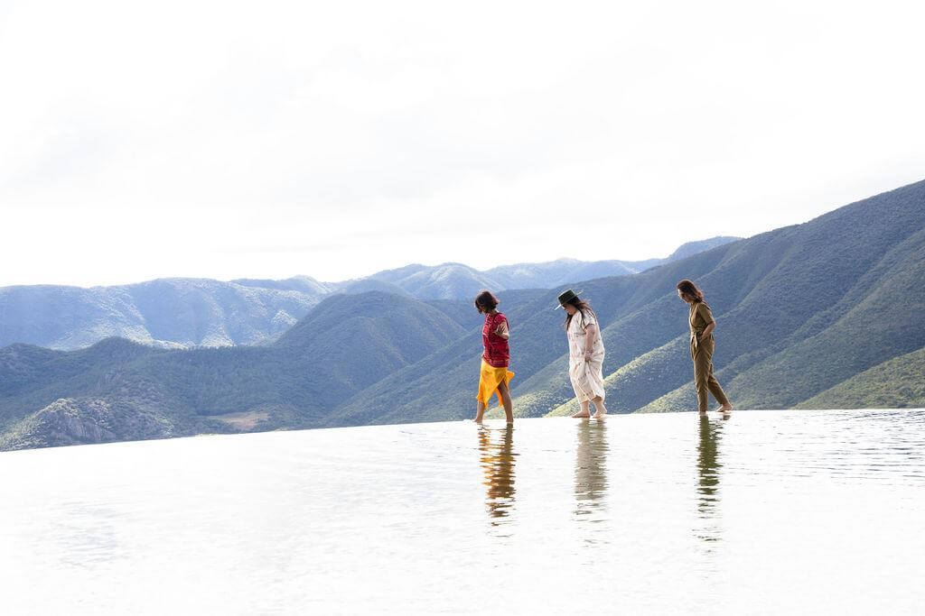 3 women walking along edge of infinity pool with tree covered hills as ackground.  one women is wearing red and orange top and skirt woman 2 a long white dress and woman 3 khaki trousers and top